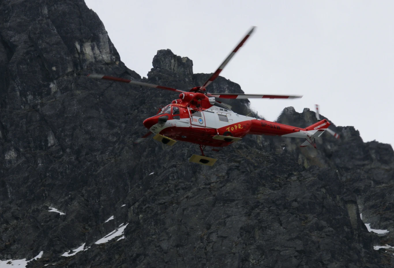 Tatrzańskie jeziora zamarzają, TPN apeluje Czerwony śmigłowiec ratunkowy leci na tle surowych, skalistych szczytów gór z widocznymi połaciami śniegu.