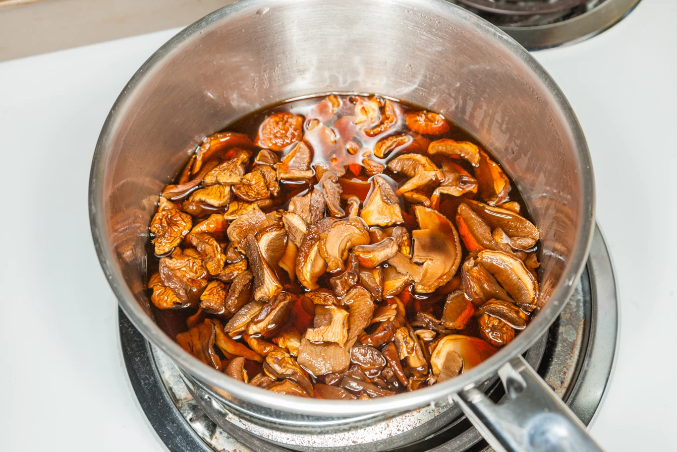 W garnku z nierdzewnej stali gotują się namoczone suszone grzyby w ciemnym wywarze. Naczynie stoi na kuchence, płyn ma intensywny brązowy kolor, a kawałki grzybów unoszą się na powierzchni wywaru.
