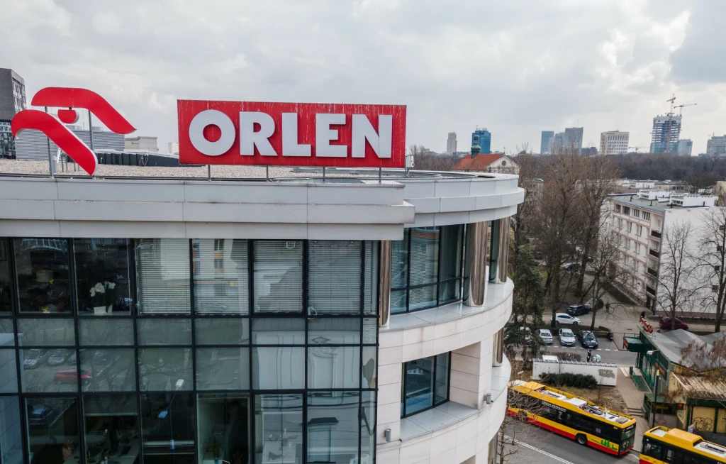 Budynek firmy Orlen z dużym czerwonym logo na dachu, w tle widoczne są drzewa, miejskie zabudowania oraz fragmenty ulicy z żółtym autobusem.