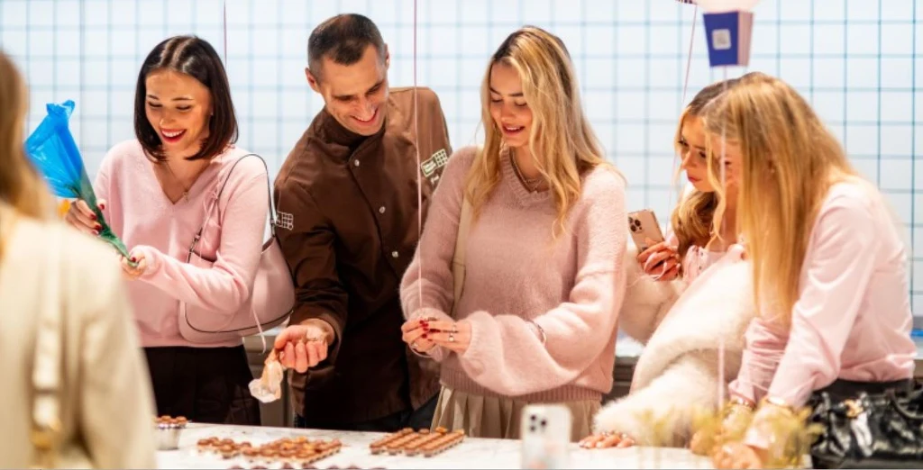 Grupa uśmiechniętych osób stoi przy stole, na którym znajdują się cukiernicze dekoracje i narzędzia do ozdabiania wyrobów, wszyscy zaangażowani są w dekorowanie słodyczy.