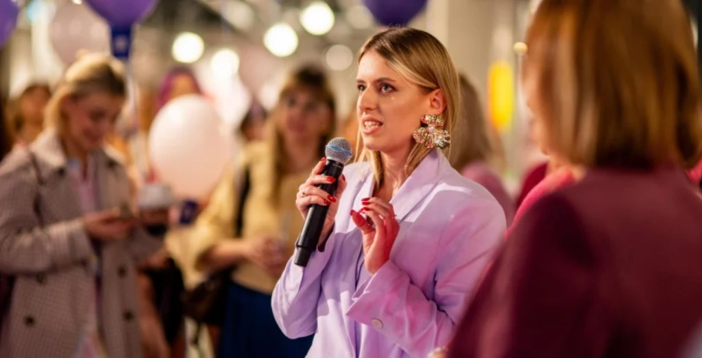 Kobieta w jasnym żakiecie trzyma mikrofon i przemawia do zgromadzonej grupy osób w tle, które słuchają lub robią zdjęcia. W pomieszczeniu panuje ciepłe, przyjazne oświetlenie, a wokół widoczna jest swobodna atmosfera spotkania lub wydarzenia.