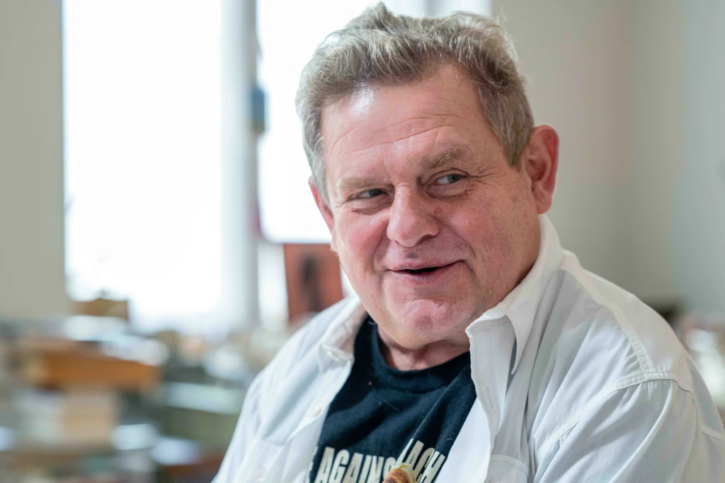 Kazik Staszewski (Kult) wydał specjalne oświadczenie Starszy mężczyzna z siwymi włosami uśmiecha się, ubrany w ciemny podkoszulek i rozpiętą białą koszulę, tło rozmyte z jasnym światłem z okna, atmosfera pozytywna i swobodna.