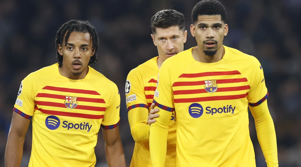 Trzech piłkarzy klubu FC Barcelona w żółtych strojach z czerwonymi paskami, widocznych na pierwszym planie na tle stadionu podczas meczu piłkarskiego.