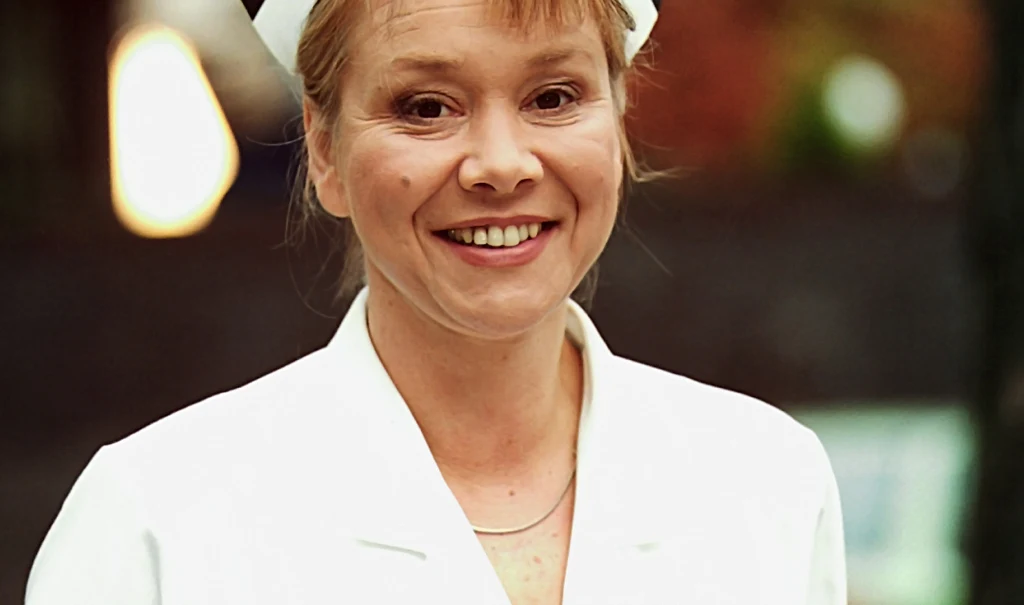 Uśmiechnięta kobieta w białym fartuchu i czepku pielęgniarskim na głowie, stojąca na zewnątrz, z rozmytym tłem w ciemnych barwach.