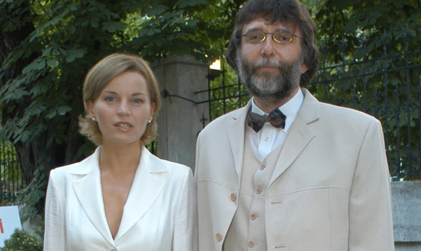 Dwie osoby ubrane elegancko stoją obok siebie na tle zieleni oraz metalowego ogrodzenia. Kobieta ma jasne włosy i jasny garnitur, mężczyzna ma brodę, okulary i jasny garnitur z muchą.