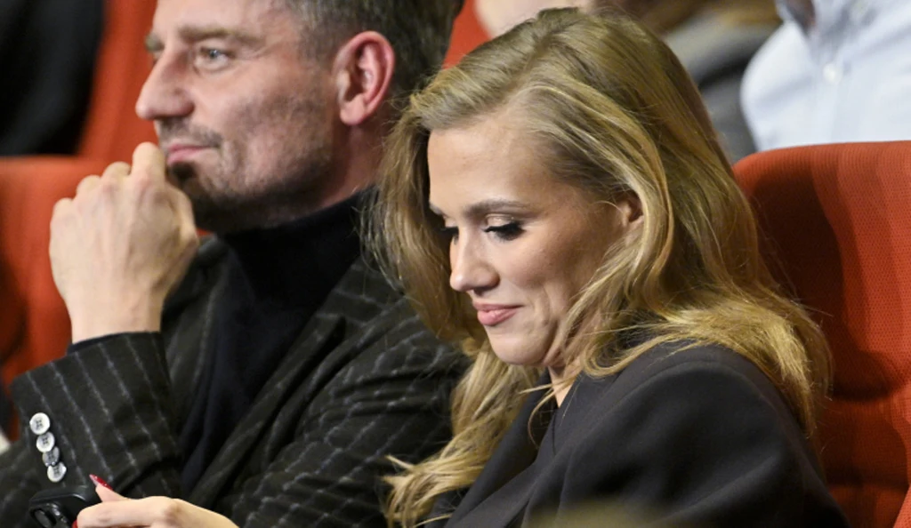 Dwoje elegancko ubranych osób siedzących obok siebie na czerwonych fotelach w sali widowiskowej, kobieta z długimi blond włosami skierowana w stronę ekranu telefonu.