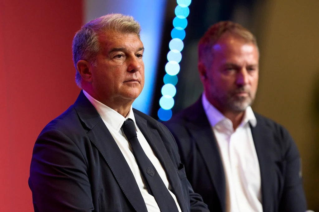 Dwóch mężczyzn w garniturach siedzi obok siebie na tle czerwonej i niebieskiej scenerii. Obaj mają poważny wyraz twarzy.