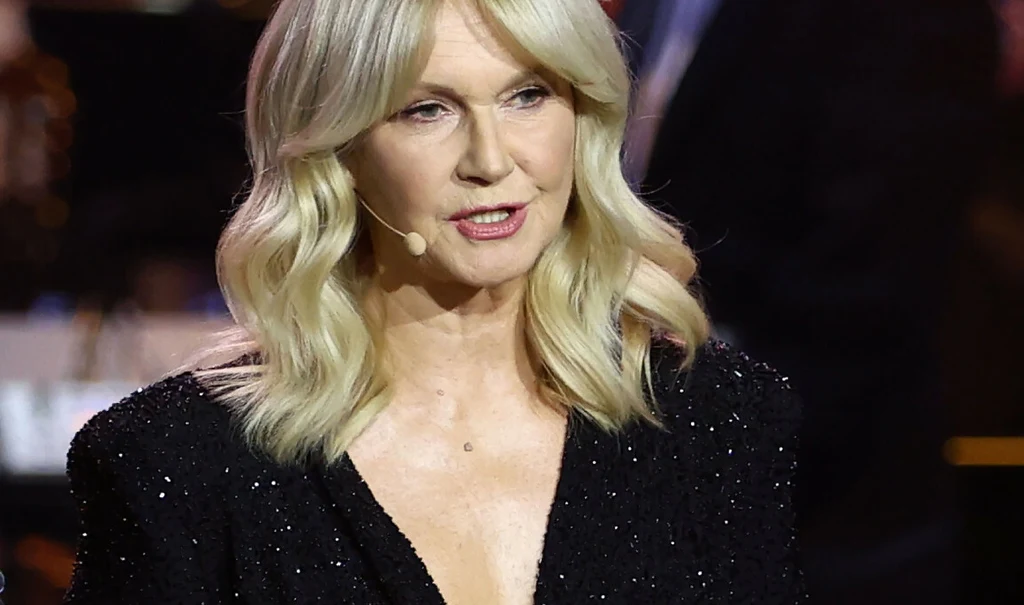 Kobieta o blond włosach występuje na scenie, ubrana w czarną, błyszczącą suknię z głębokim dekoltem, z mikrofonem nagłownym, w tle widoczne rozmyte elementy orkiestry lub zespołu muzycznego.