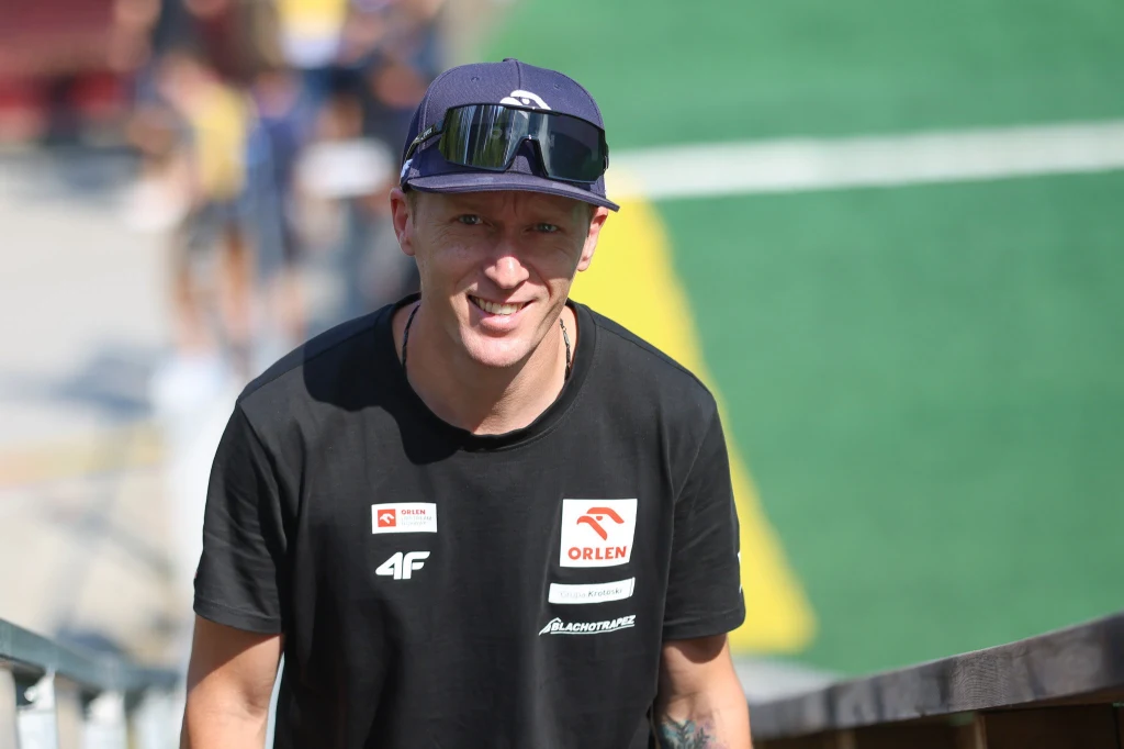 Mężczyzna w sportowej koszulce z logotypami sponsorskimi oraz ciemnej czapce i okularach przeciwsłonecznych stoi na tle zielonego boiska, uśmiecha się i spogląda w kierunku aparatu.