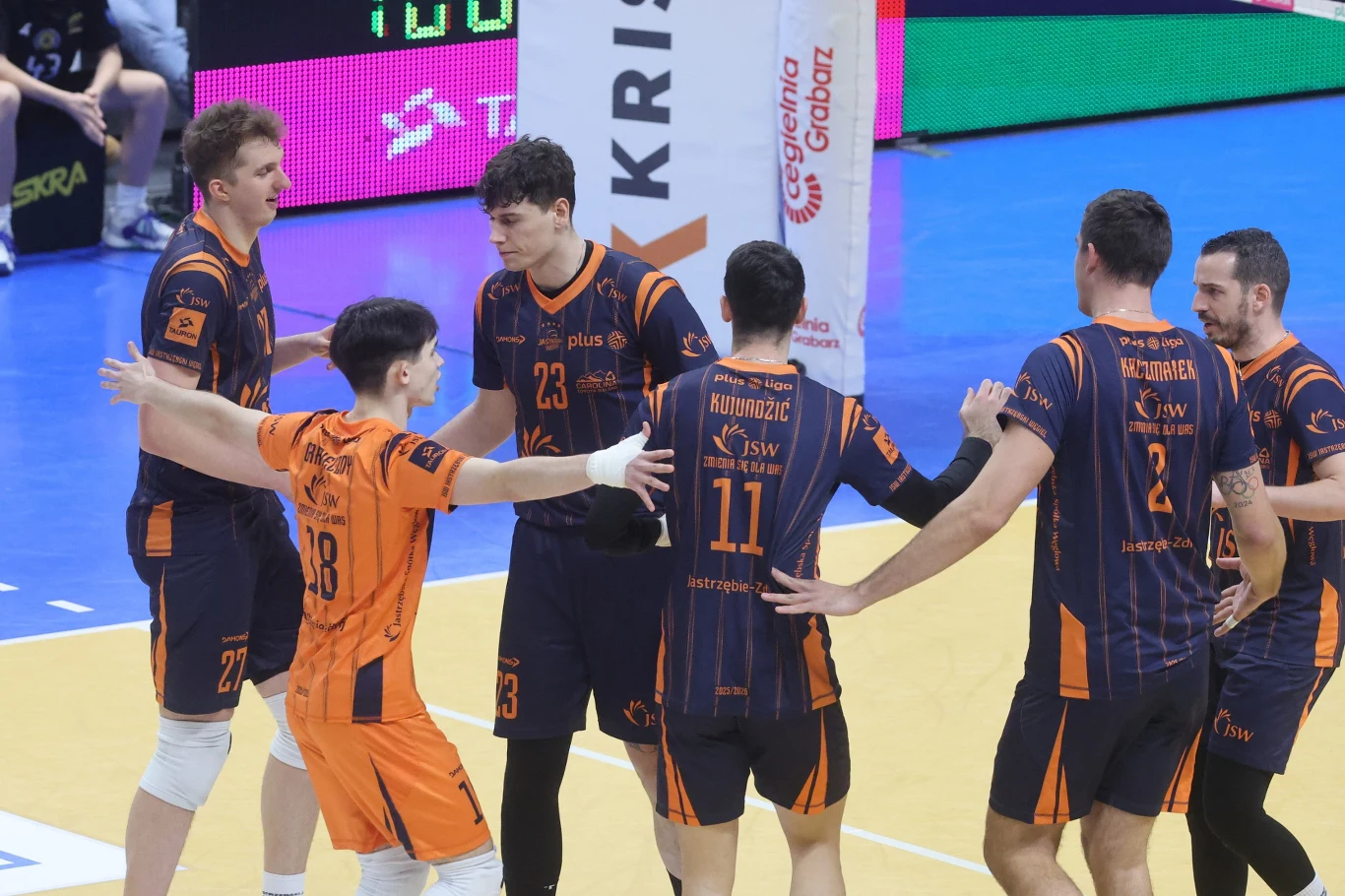 Siatkarze JSW Jastrzębskiego Węgla Zawodnicy drużyn siatkarskich w trakcie meczu na hali, w wyrazistych pomarańczowych i granatowych strojach sportowych podczas sportowej interakcji na parkiecie.