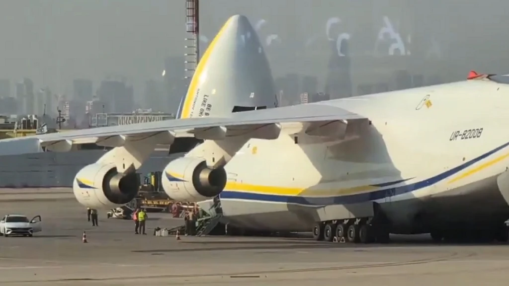 Największy na świecie samolot transportowy Antonow An-225 Mrija stojący na płycie lotniska, z podniesioną częścią dziobową umożliwiającą załadunek, w otoczeniu kilku osób oraz pojazdu serwisowego, w tle majaczy panorama miasta.