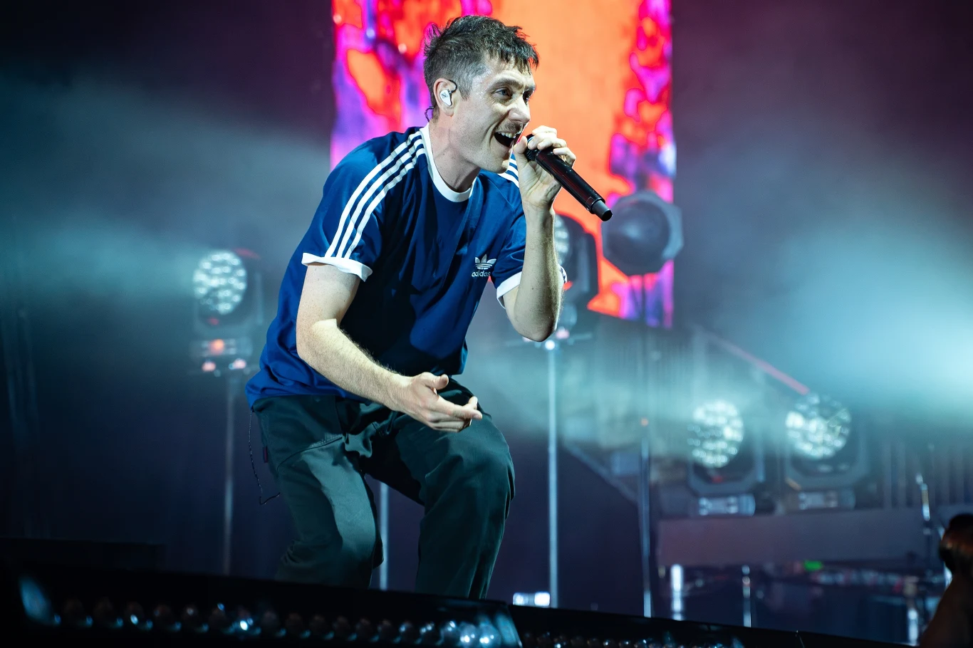 Dan Smith (Bastille) w akcji Wokalista ubrany w niebieską koszulkę Adidas śpiewa do mikrofonu na scenie koncertowej, oświetlony kolorowym światłem z efektami wizualnymi w tle.