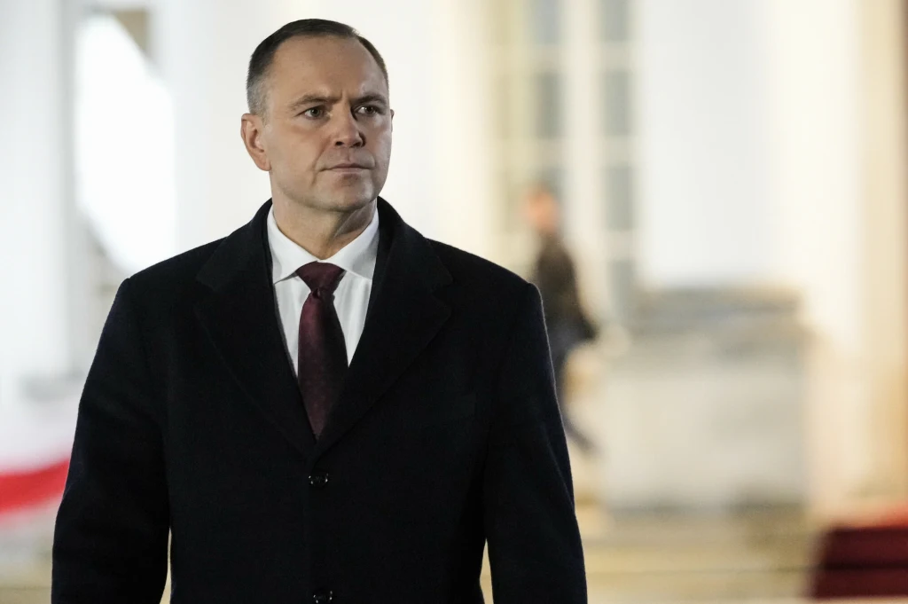 Mężczyzna w eleganckim, ciemnym płaszczu i bordowym krawacie stoi na tle jasnego budynku z kolumnami, w tle widoczne są rozmazane sylwetki osób.