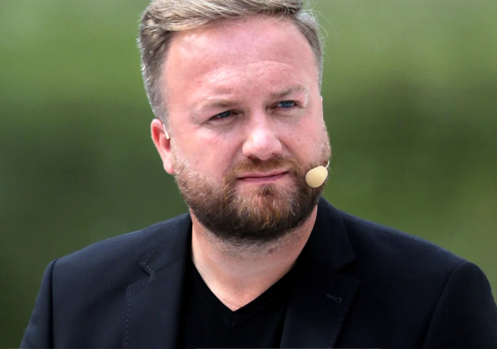 Bartłomiej Kasprzykowski Mężczyzna z jasną brodą oraz krótkimi włosami ubrany w czarną marynarkę, z mikrofonem przy twarzy, na tle rozmytego zielonego otoczenia.