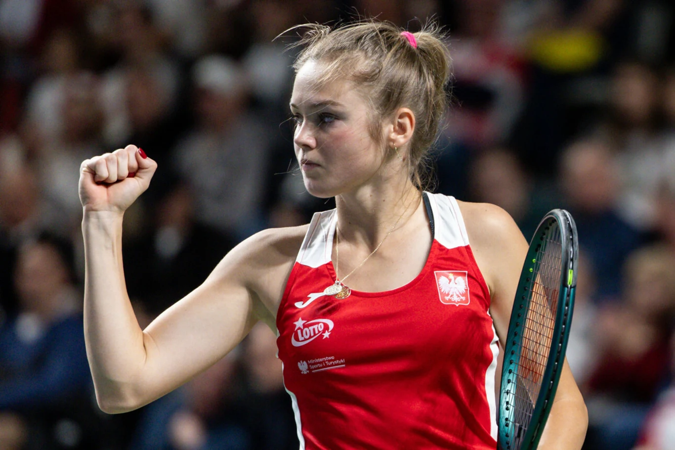 Tenisistka w czerwonym stroju sportowym wykonuje gest triumfu na korcie, trzymając rakietę tenisową. W tle widać rozmyte sylwetki kibiców.