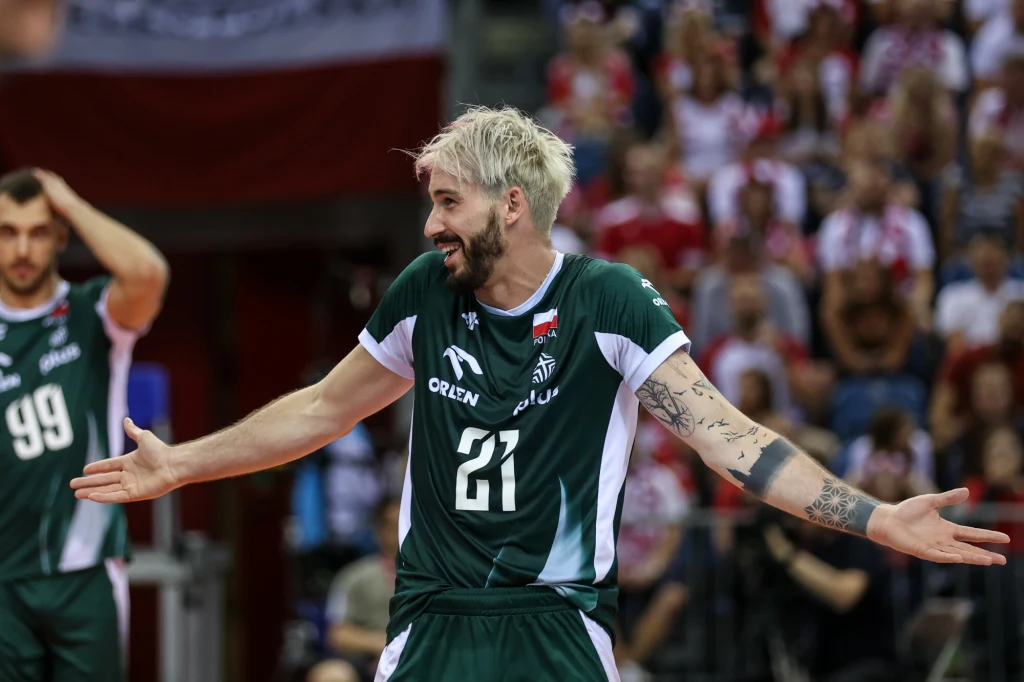 Siatkarz w zielonym stroju sportowym z widocznymi tatuażami na rękach, numer 21, stojący z szeroko rozłożonymi rękami na hali sportowej podczas meczu, w tle rozmyty tłum kibiców.