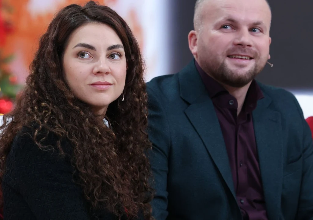 Dwoje ludzi siedzi obok siebie, kobieta z długimi, kręconymi włosami patrzy w bok, mężczyzna w marynarce uśmiecha się lekko, w tle rozmyte kolory sugerujące przyjemną atmosferę.