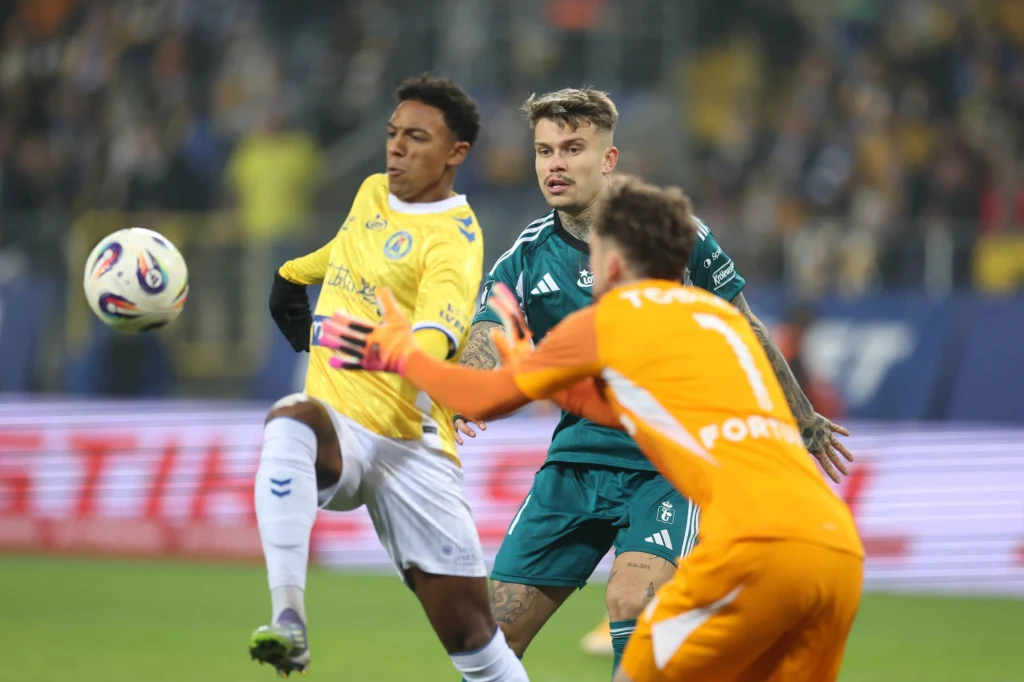 Piłkarz w żółtej koszulce kontroluje piłkę, obok znajduje się zawodnik w zielonej koszulce oraz bramkarz w pomarańczowym stroju, wszyscy intensywnie skupieni podczas meczu piłki nożnej, a w tle zamazana publiczność na trybunach.