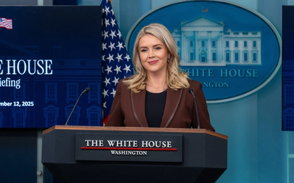 Kobieta w blond włosach stoi za mównicą w sali briefingowej Białego Domu, w tle amerykańska flaga i herb Białego Domu, profesjonalne oświetlenie oraz ekran z napisem The White House Briefing.