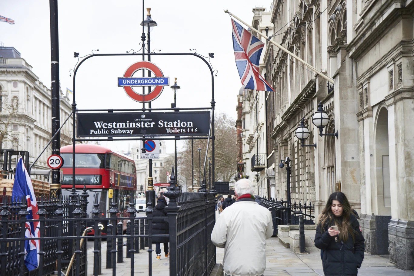 Coraz więcej Polaków decyduje się na powrót z Wysp Brytyjskich do kraju. Na zdj. Londyn Wejście do stacji metra Westminster w Londynie, widoczny znak London Underground, brytyjska flaga, zabytkowe budynki oraz przechodnie na chodniku. W tle czerwony autobus piętrowy typowy dla Londynu.