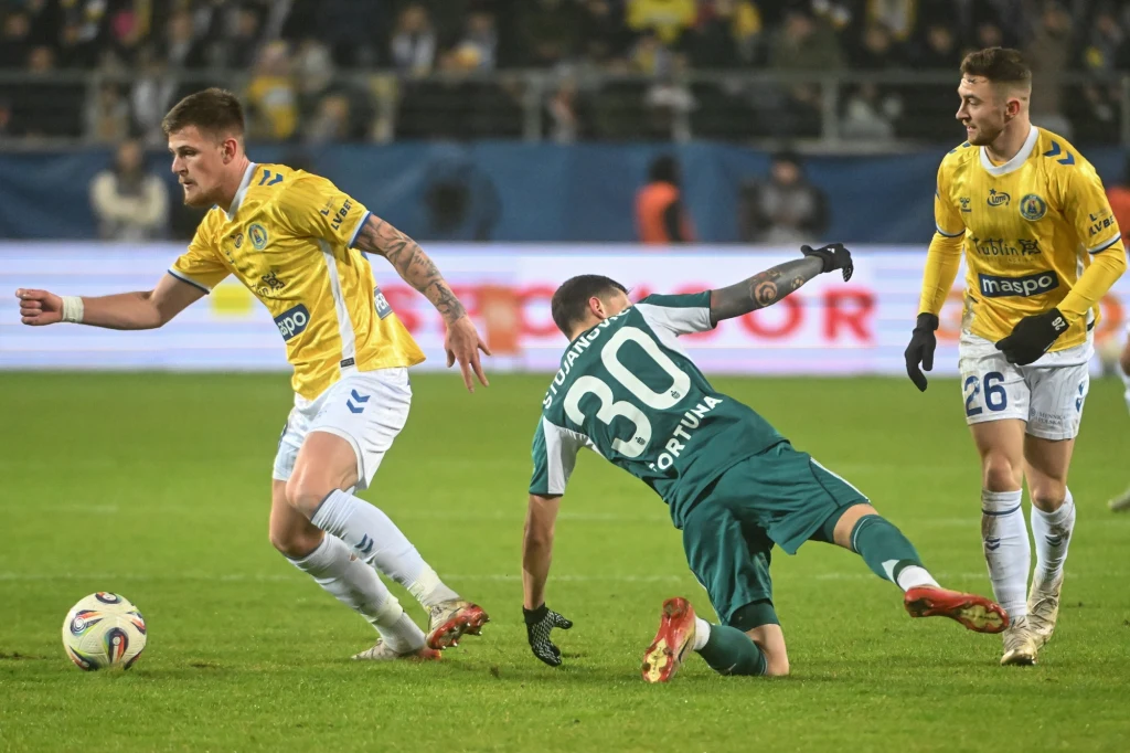 Dwóch piłkarzy w żółtych koszulkach i jeden w zielonej podczas dynamicznej akcji na boisku, jeden z zawodników upada na trawę, za nimi trybuny z kibicami.