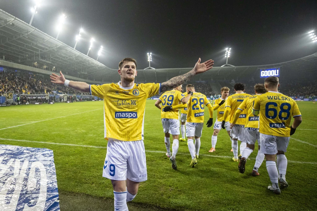 Piłkarz drużyny w żółtych koszulkach rozpościera ręce w geście radości, za nim kilku innych zawodników kieruje się w stronę kibiców na pełnym trybun stadionie, całość oświetlona jasnymi reflektorami.