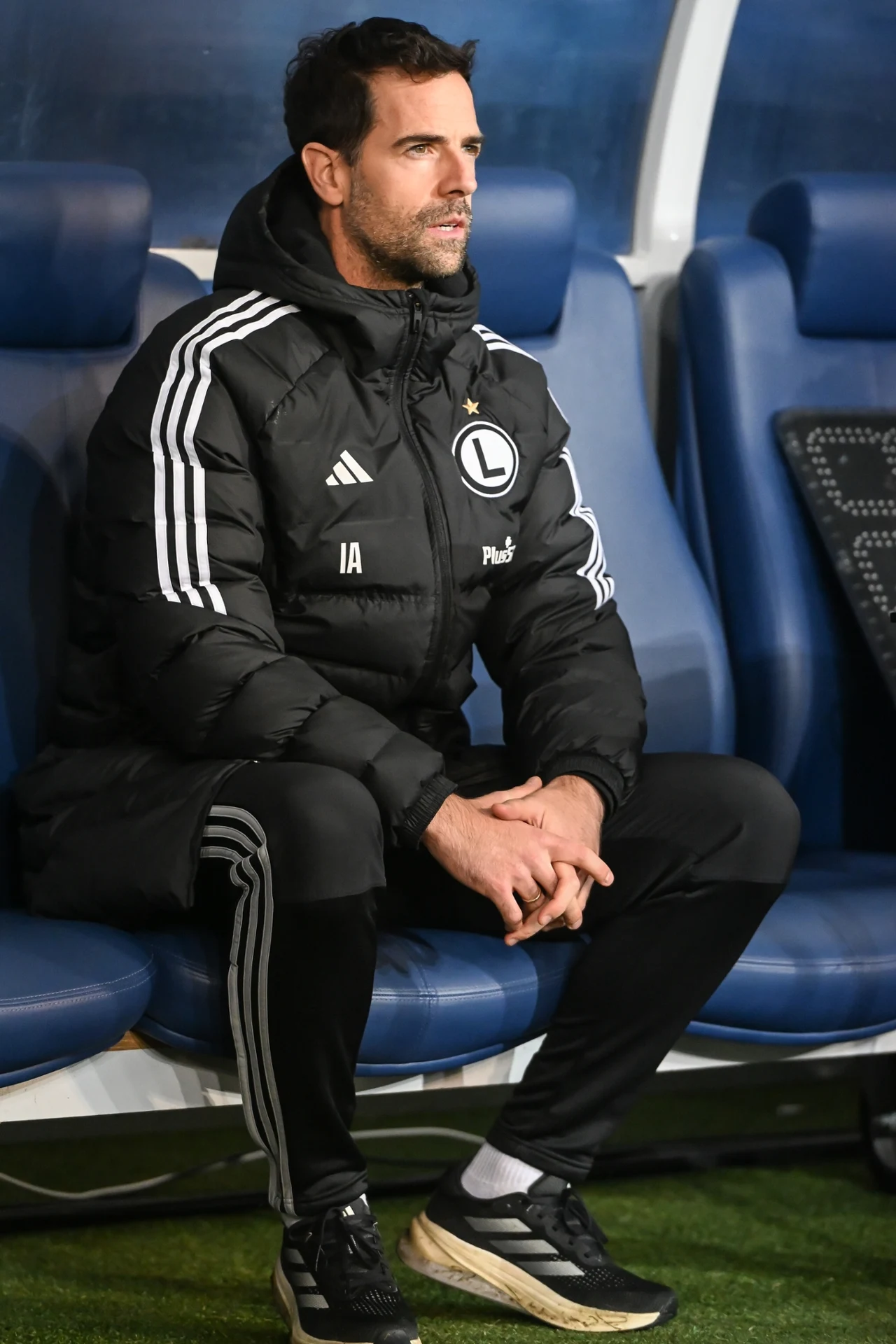 Mężczyzna w sportowej kurtce z logo Legii Warszawa siedzi na niebieskiej ławce stadionowej, z dłońmi splecionymi na kolanach.