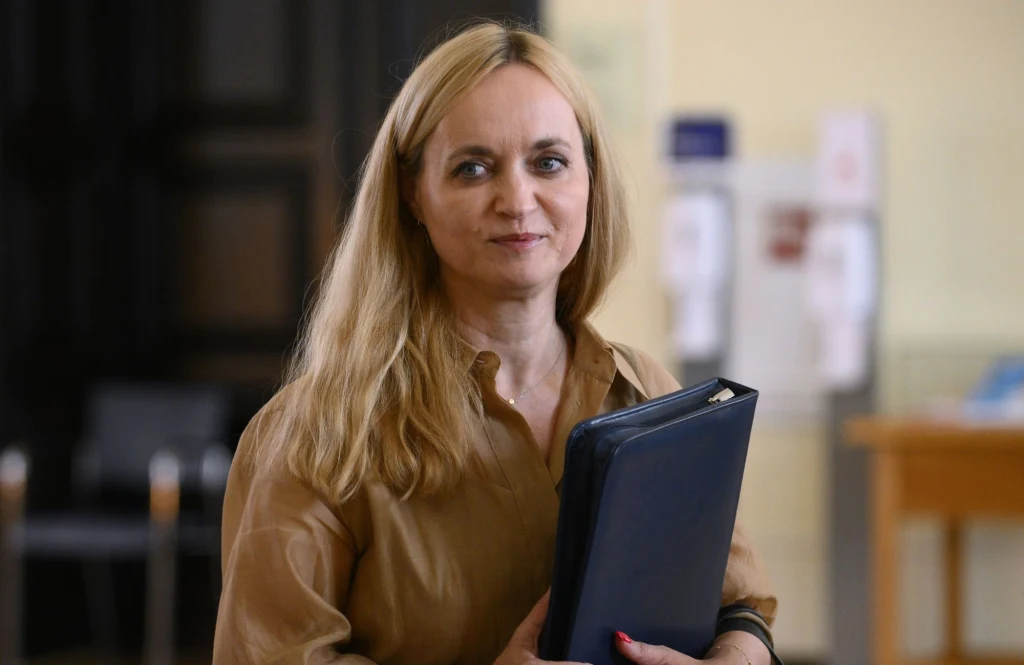 Minister zdrowia Jolanta Sobierańska-Grenda Kobieta z długimi blond włosami ubrana w jasnobrązową koszulę trzyma granatową teczkę w dłoni. Uśmiecha się lekko, stoi w jasnym pomieszczeniu z rozmazanym tłem, gdzie widać fragment krzesła i mebli.