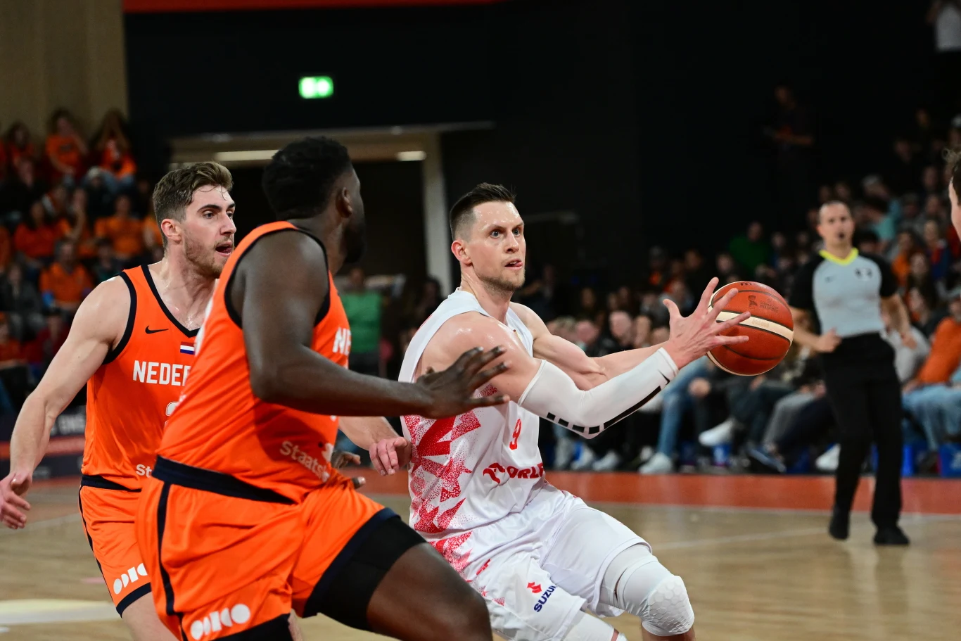 Mateusz Ponitka, kapitan reprezentacji Polski Koszykarz w białym stroju próbuje zdobyć punkt, dynamicznie atakując kosz pomiędzy dwoma zawodnikami w pomarańczowych strojach reprezentacji Holandii, na tle widoczna publiczność i sędzia.