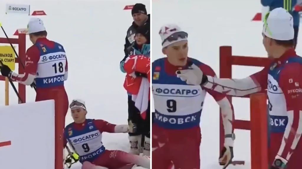 Dwóch biegaczy narciarskich w czerwonych strojach, jeden siedzi wyczerpany na śniegu, drugi gestykuluje ręką w jego stronę. W tle kilku kibiców oraz tablice oznaczeniowe.
