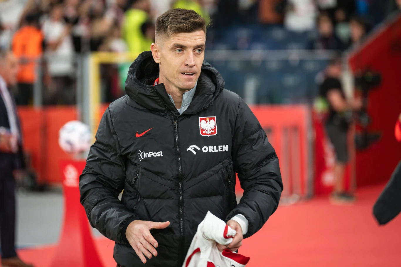 Krzysztof Piątek Mężczyzna w sportowej kurtce z emblematem polskiej reprezentacji piłkarskiej, trzymający sportową koszulkę w dłoniach, stoi na stadionie z rozmytym tłumem oraz elementami boiska w tle.