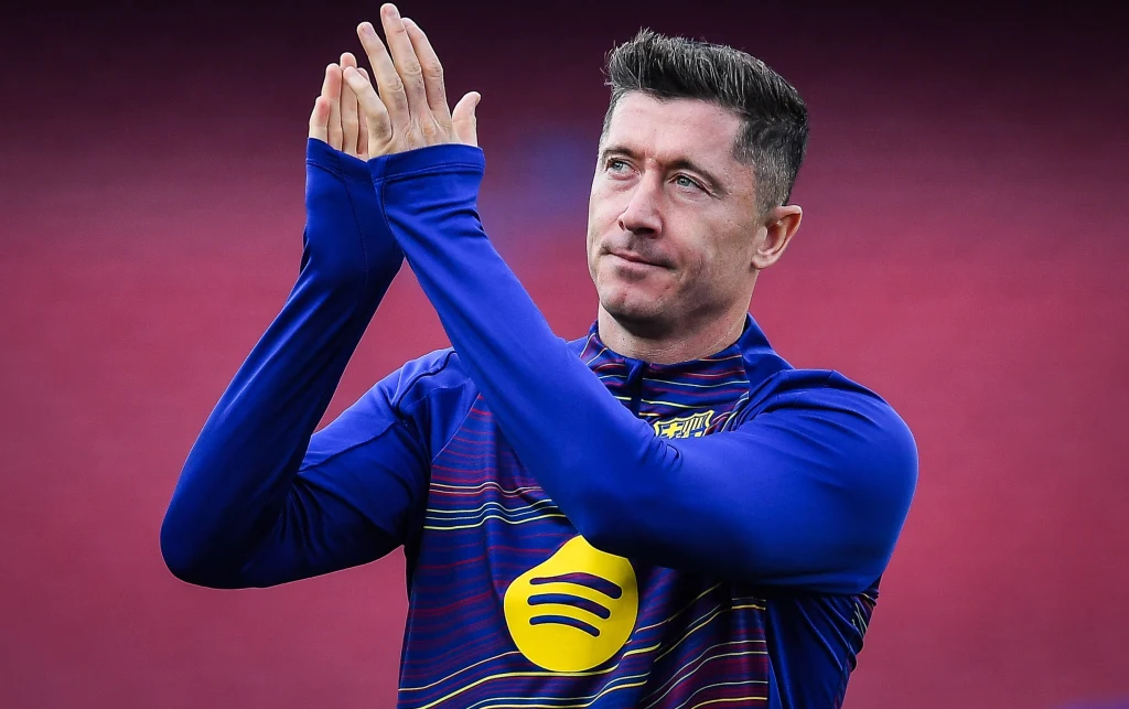 Robert Lewandowski Piłkarz w sportowej koszulce klubu piłkarskiego z logo sponsora na piersi, wykonujący gest składania dłoni jakby bił brawo, na jednolitym, ciemnym tle.