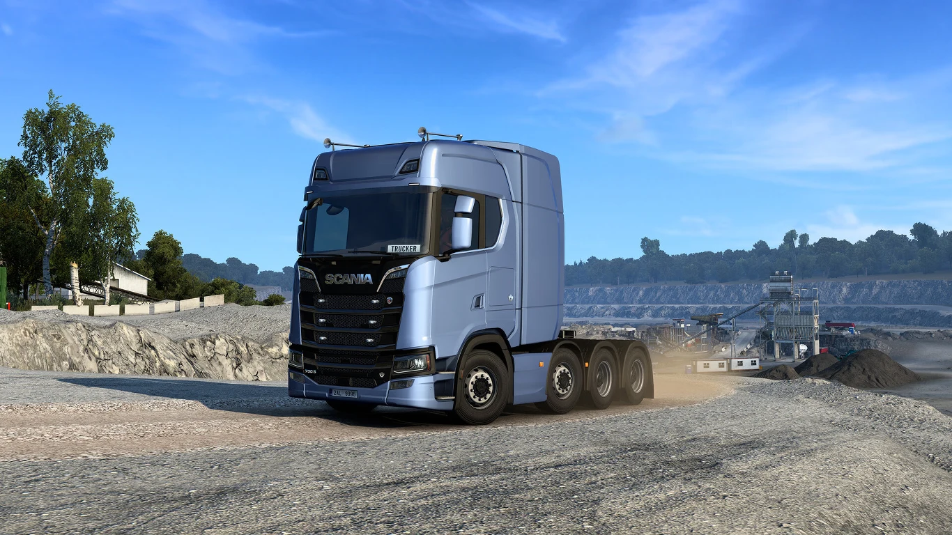 Euro Truck Simulator Srebrna ciężarówka marki Scania jedzie po piaszczystej drodze przez industrialny krajobraz kopalni, w tle widoczne maszyny wydobywcze oraz niska zabudowa po lewej stronie.