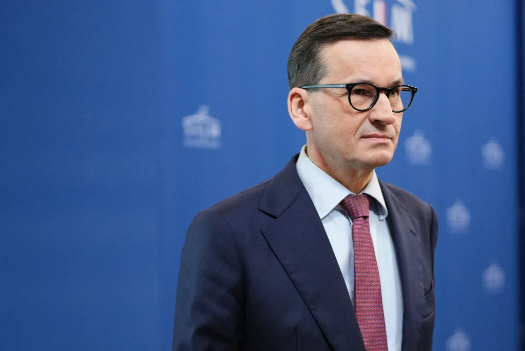 "Podatek Morawieckiego" wciąż nie zniknął. Oto plan rządu Mężczyzna w garniturze i okularach stoi na tle niebieskiej ściany z rozmytym logo w tle. Ma poważny wyraz twarzy, jest dobrze ubrany, a jego sylwetka dominuje na pierwszym planie.