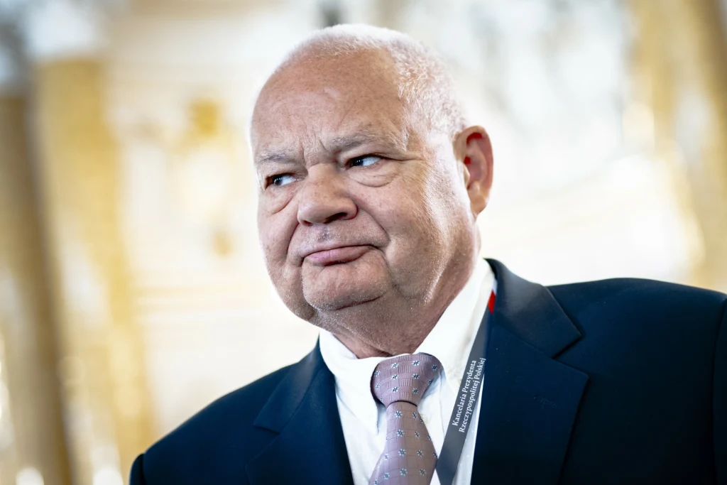 RPP obniży stopy procentowe podczas grudniowego posiedzenia? Na zdj. prezes NBP i przewodniczący RPP Adam Glapiński Starszy mężczyzna ubrany w ciemny garnitur, białą koszulę i krawat, z identyfikatorem na szyi, znajduje się w jasnym, eleganckim wnętrzu.