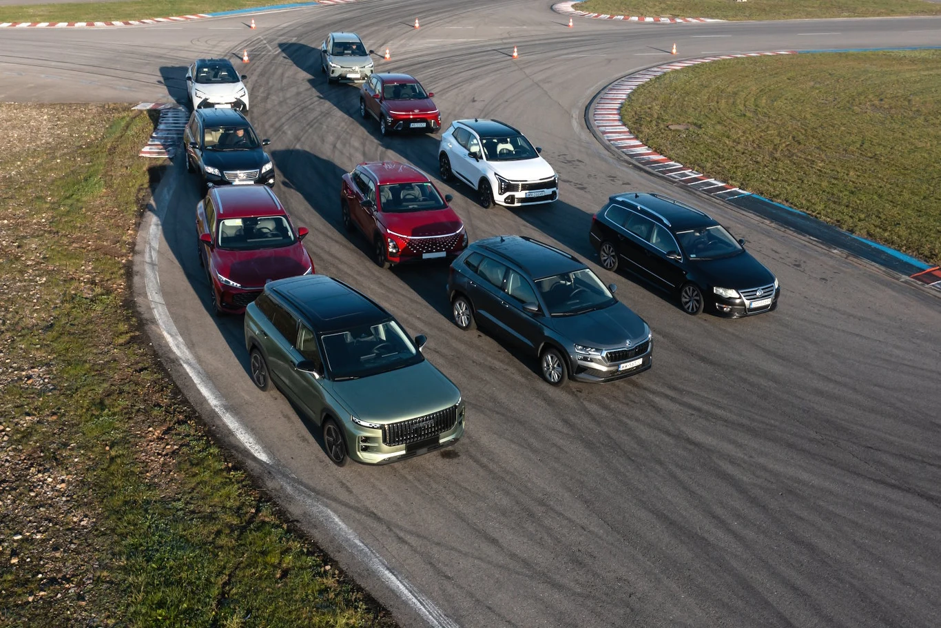 Stary Volkswagen Passat B6 TDI czy nowe chińskie SUVy? Które auto lepiej radzi sobie na torze? Grupa nowoczesnych samochodów osobowych różnych marek ustawiona równo na torze wyścigowym, widoczny asfalt toru, barwna karoseria pojazdów oraz lekko zakręcająca nawierzchnia, a w tle fragment zielonego terenu.