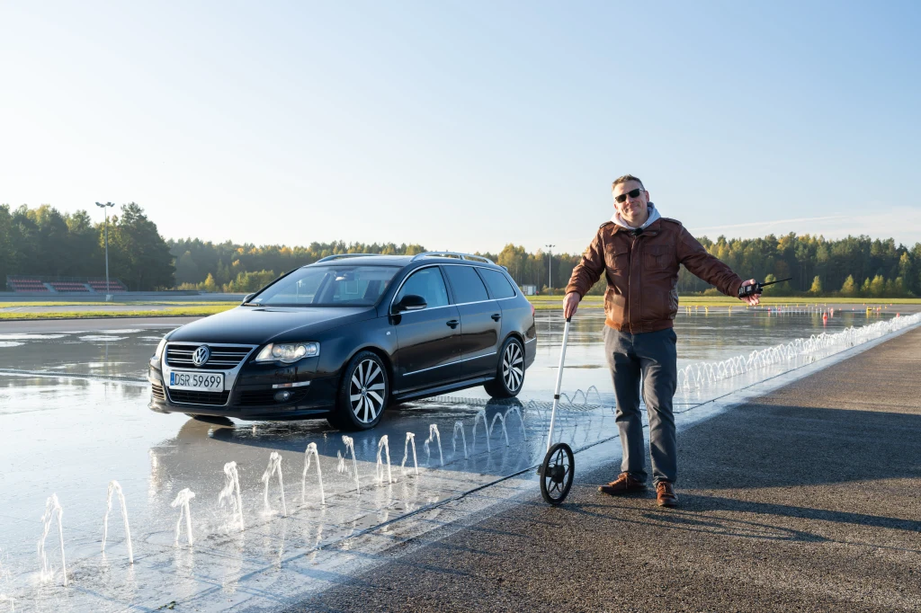 Mężczyzna w brązowej kurtce i okularach przeciwsłonecznych stoi obok miernika pomiarowego na torze testowym, na którym znajduje się ciemny samochód osobowy Volkswagen Passat, w pobliżu ustawione są rozpryski wodne symulujące trudne warunki jazdy