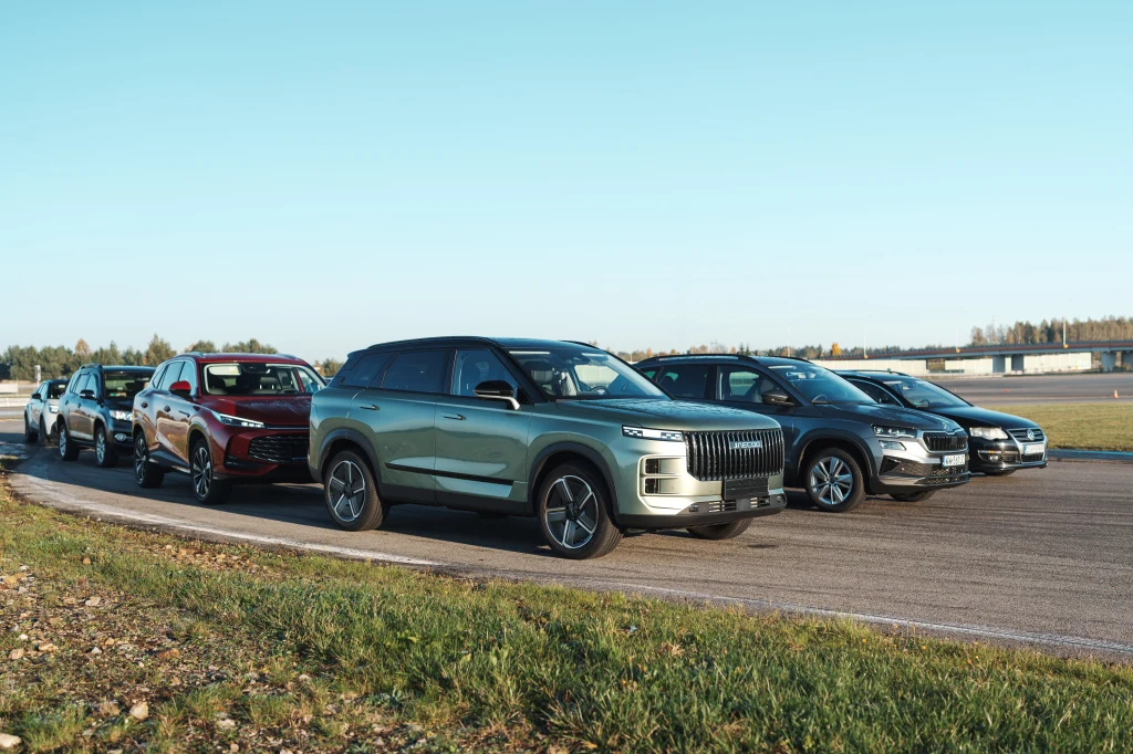 Sześć nowoczesnych samochodów osobowych ustawionych w rzędzie na torze testowym, w otoczeniu zieleni i asfaltowej nawierzchni, przy jasnym, słonecznym niebie.