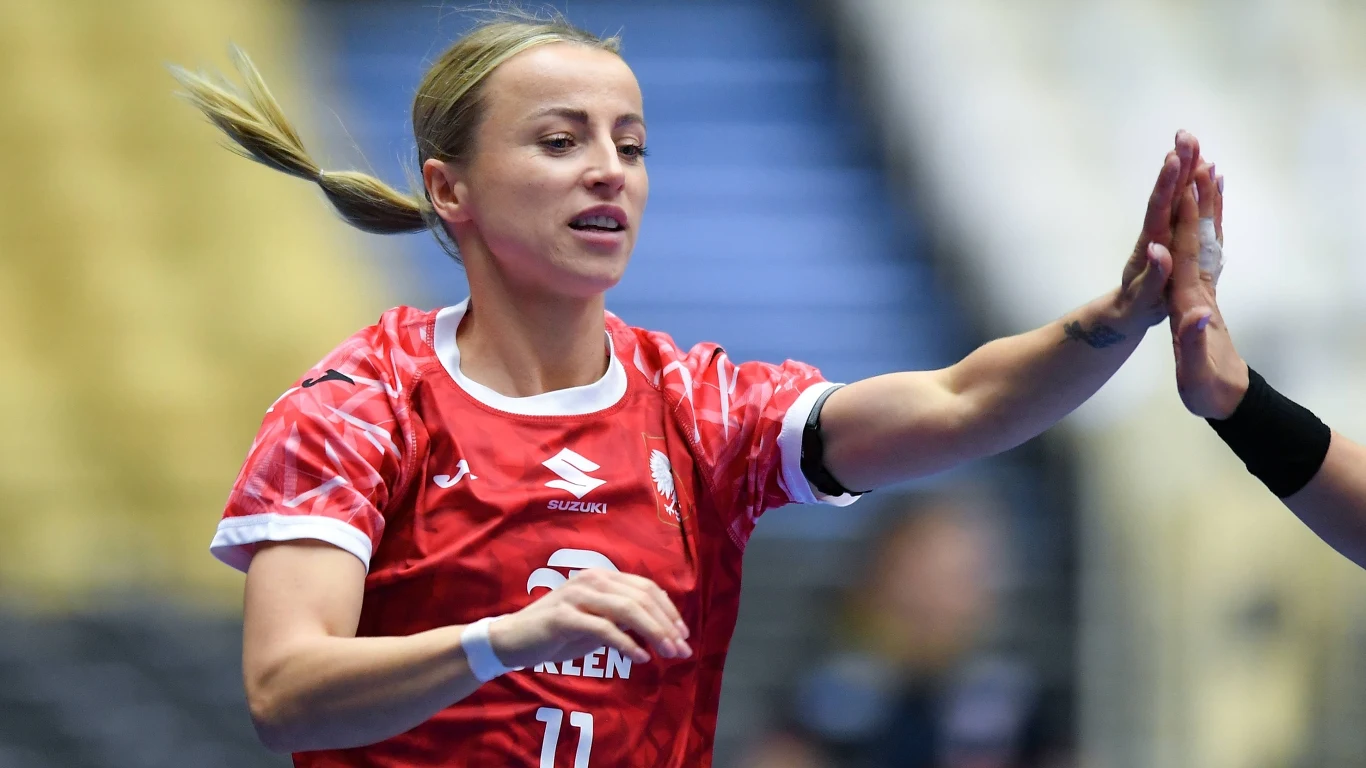 Mariola Wiertelak Zawodniczka w czerwonym stroju sportowym z numerem 11 wykonuje gest przybicia piątki z inną osobą na tle rozmytej hali sportowej.