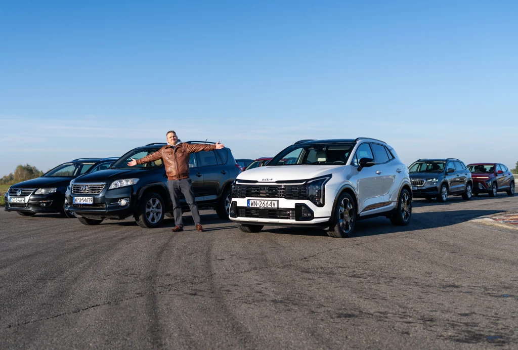 Grupa nowoczesnych samochodów osobowych ustawionych na asfalcie, z mężczyzną w skórzanej kurtce stojącym z rozłożonymi ramionami przed pojazdami, pogodny dzień i czyste niebo w tle.