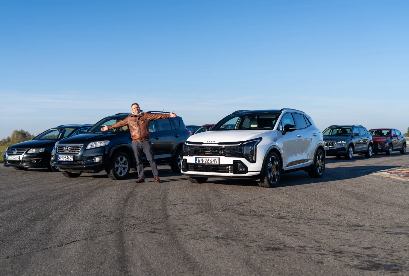 Nowa Kia Sportage miała na torze spore problemy. W próbach na mokrej nawierzchni lepiej radziły sobie stary Volkswagen Passat i Toyota RAV4 Grupa nowoczesnych samochodów osobowych ustawionych na asfalcie, z mężczyzną w skórzanej kurtce stojącym z rozłożonymi ramionami przed pojazdami, pogodny dzień i czyste niebo w tle.