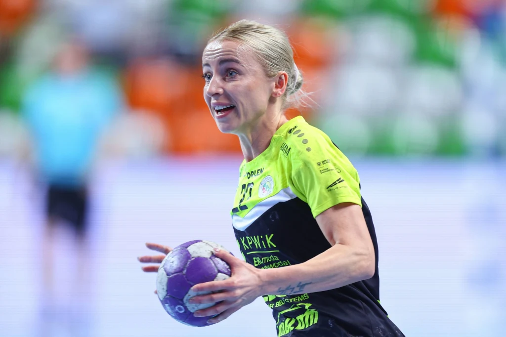 Zawodniczka piłki ręcznej w jasnozielono-czarnym stroju trzyma piłkę w dłoniach, wygląda na skupioną i dynamiczną, w tle rozmyte kolory trybun i sylwetka innej osoby.