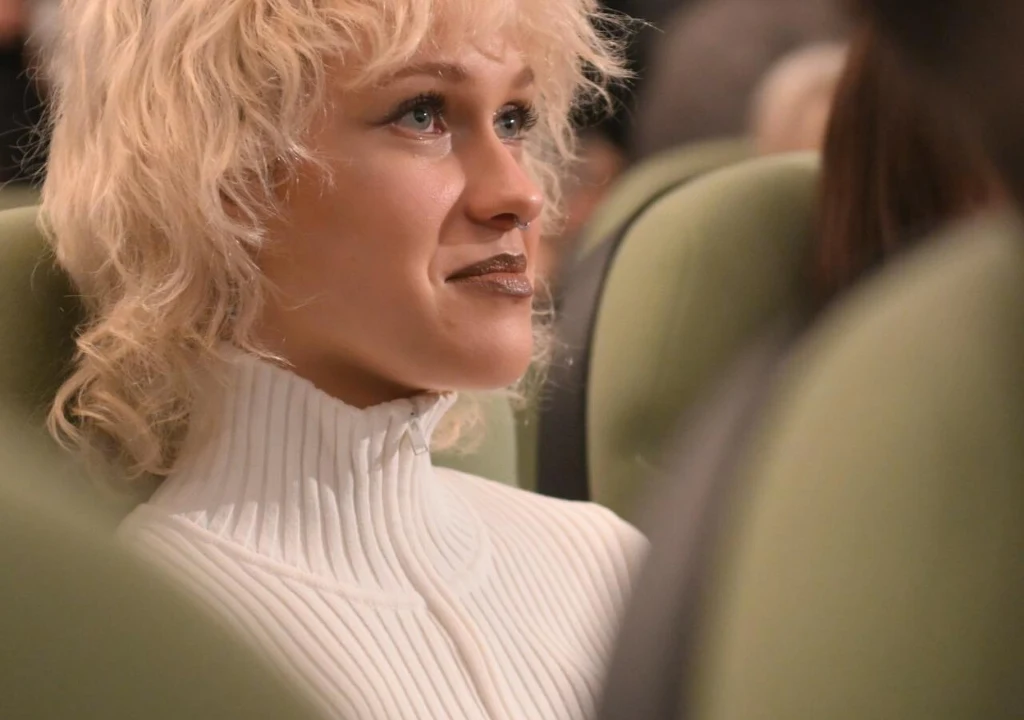 Młoda kobieta o jasnych, kręconych włosach siedzi na jasnozielonym fotelu w garniturze teatralnym lub kinowym, ubrana w biały golf, patrzy z lekkim uśmiechem w górę.