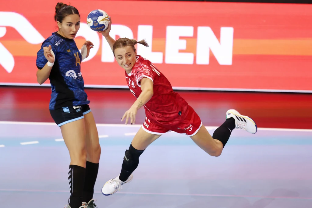 Zawodniczka w czerwonym stroju rzuca piłkę ręczną, będąc w wyskoku, obok znajduje się rywalka ubrana w niebieski strój sportowy. W tle widoczna jest część boiska i reklama z napisem 'Orlen'.