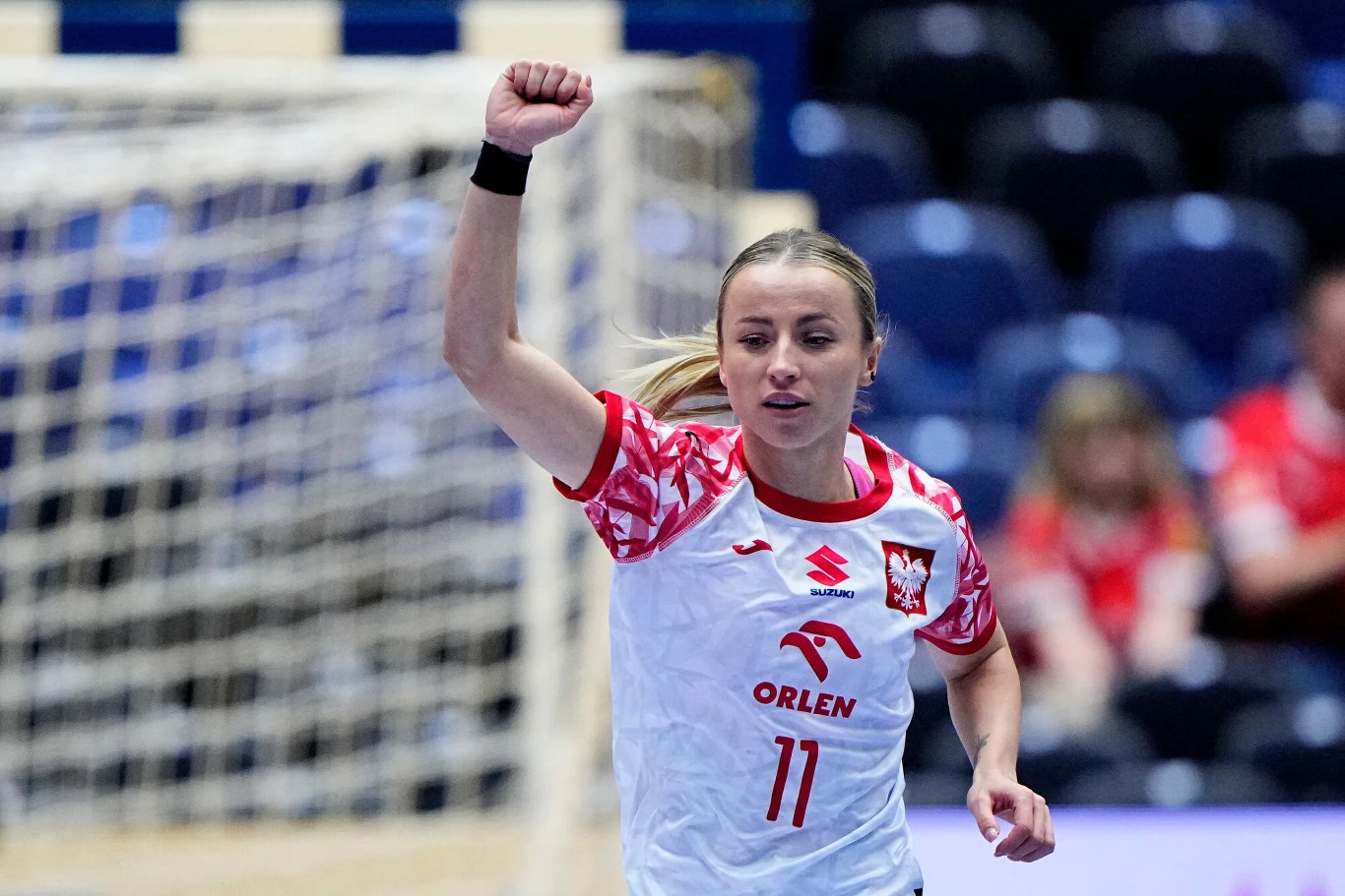 Mariola Wiertelak Zawodniczka piłki ręcznej w białej koszulce z numerem 11 unosi zaciśniętą pięść w geście triumfu na tle bramki i trybun.