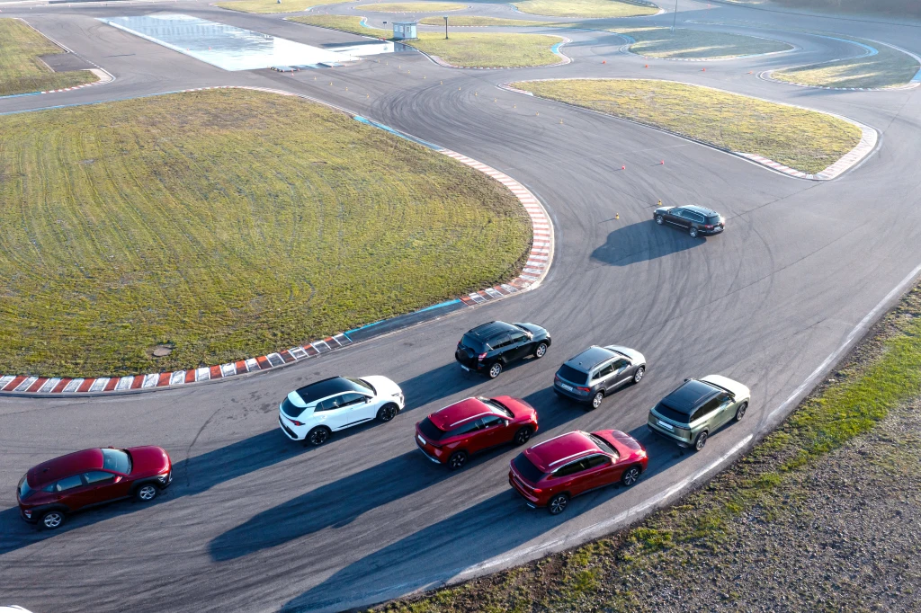 Siedem samochodów osobowych ustawionych w rzędzie na asfaltowym torze wyścigowym, otoczonym trawiastymi fragmentami oraz zakrętami toru, w słoneczny dzień.