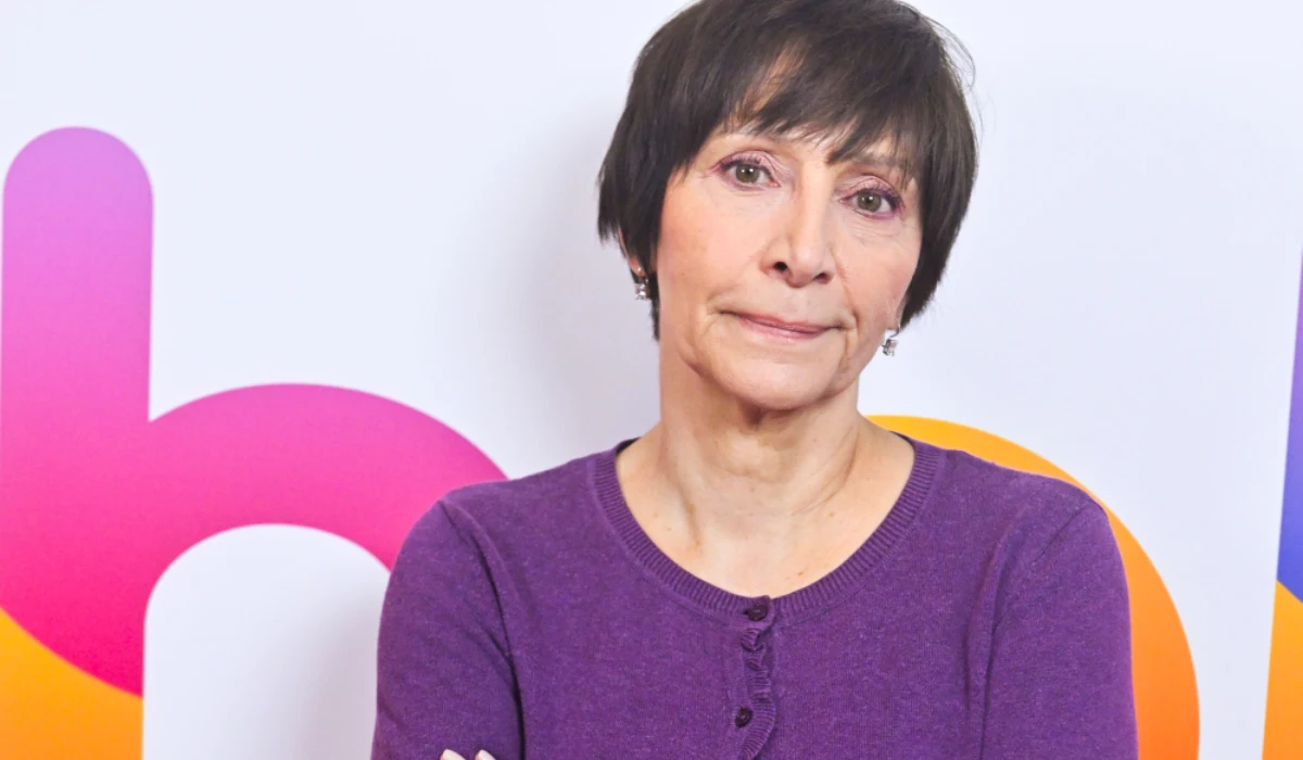 Renata Pałys Kobieta w średnim wieku z krótkimi, ciemnymi włosami ubrana w fioletowy sweter stoi na tle kolorowej grafiki z wyraźnymi literami i elementami w barwach pomarańczowych, różowych i fioletowych.