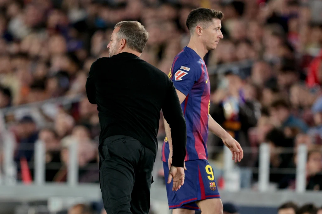 Piłkarz w stroju FC Barcelony z numerem 9 na plecach idzie po boisku, obok niego znajduje się mężczyzna w czarnym ubraniu, w tle rozmazany tłum kibiców na trybunach.