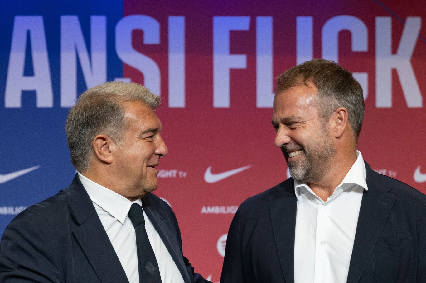 Dwóch elegancko ubranych mężczyzn w garniturach uśmiecha się do siebie, stojąc na tle czerwono-niebieskiego baneru z widocznym napisem i logotypami Nike.