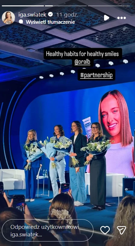 Cztery kobiety stojące na scenie, każda trzyma bukiet kwiatów. W tle widoczny jest duży ekran z wizerunkiem jednej z obecnych kobiet uśmiechającej się i trzymającej pastę do zębów. Dookoła panuje oficjalna i profesjonalna atmosfera eventu.